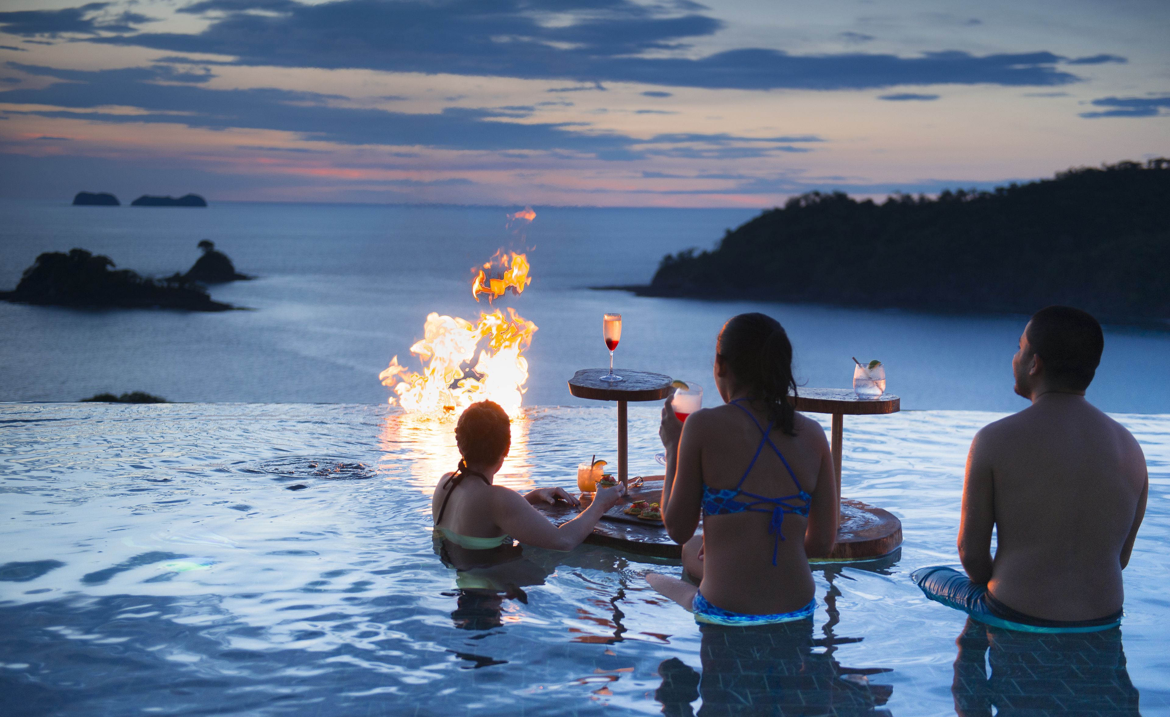 A poolside setting with a fire feature, a table with drinks, and a view of the sea and distant land.