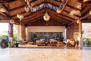 A spacious indoor area with a high ceiling, wooden beams, and hanging pendant lights. There are seating arrangements and a wall with shelves displaying various items. - Casa Chameleon Hotel Las Catalinas - Adults Only (Tempate)