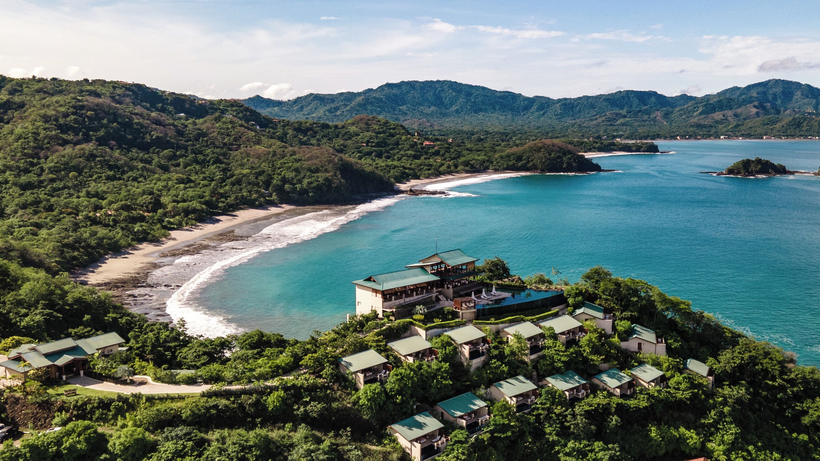 Un complejo turístico en la costa con varias edificaciones, una playa y montañas al fondo.