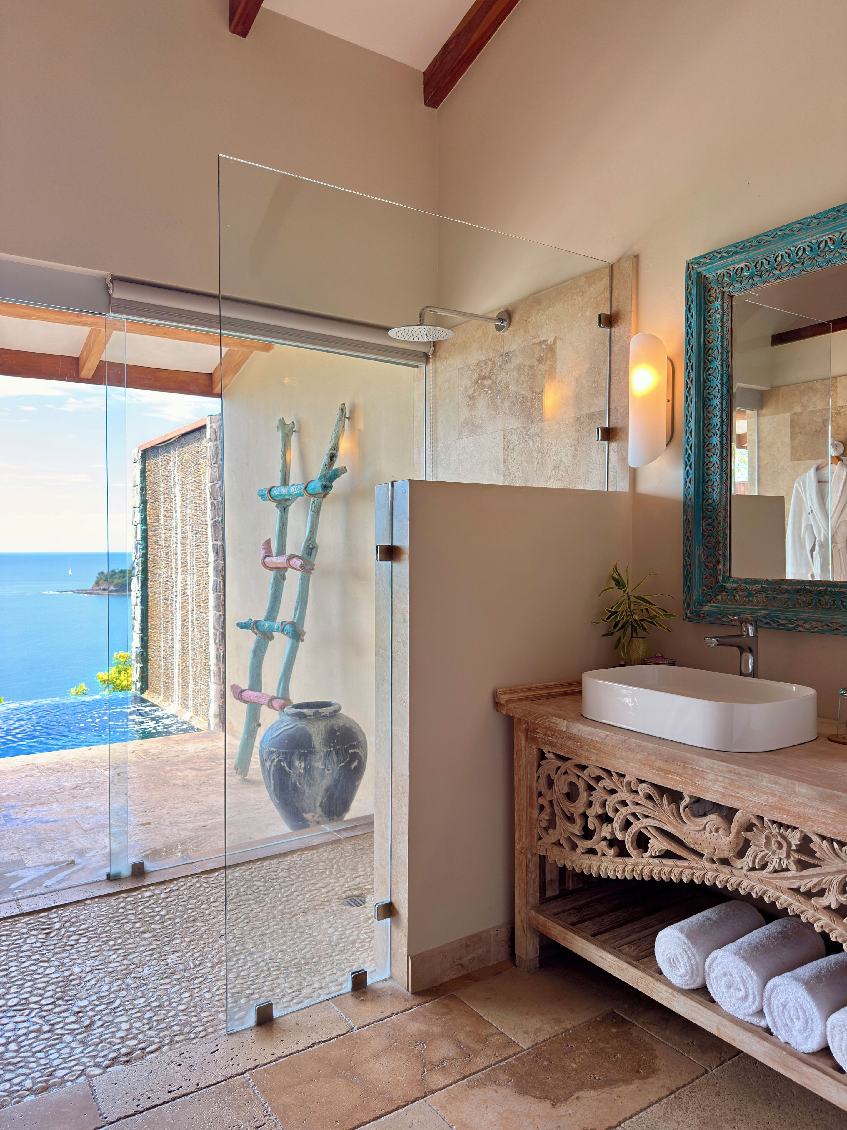 A bathroom with a large mirror, a sink, and a shower. There is a view of the sea through a glass door.