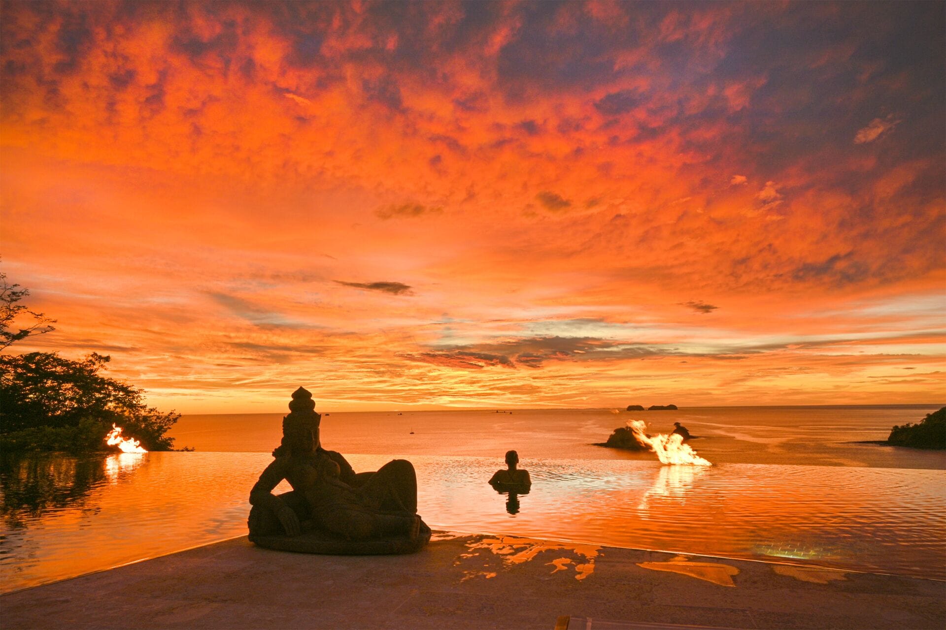 Un coucher de soleil sur une mer calme, avec une statue et une personne dans l’eau.