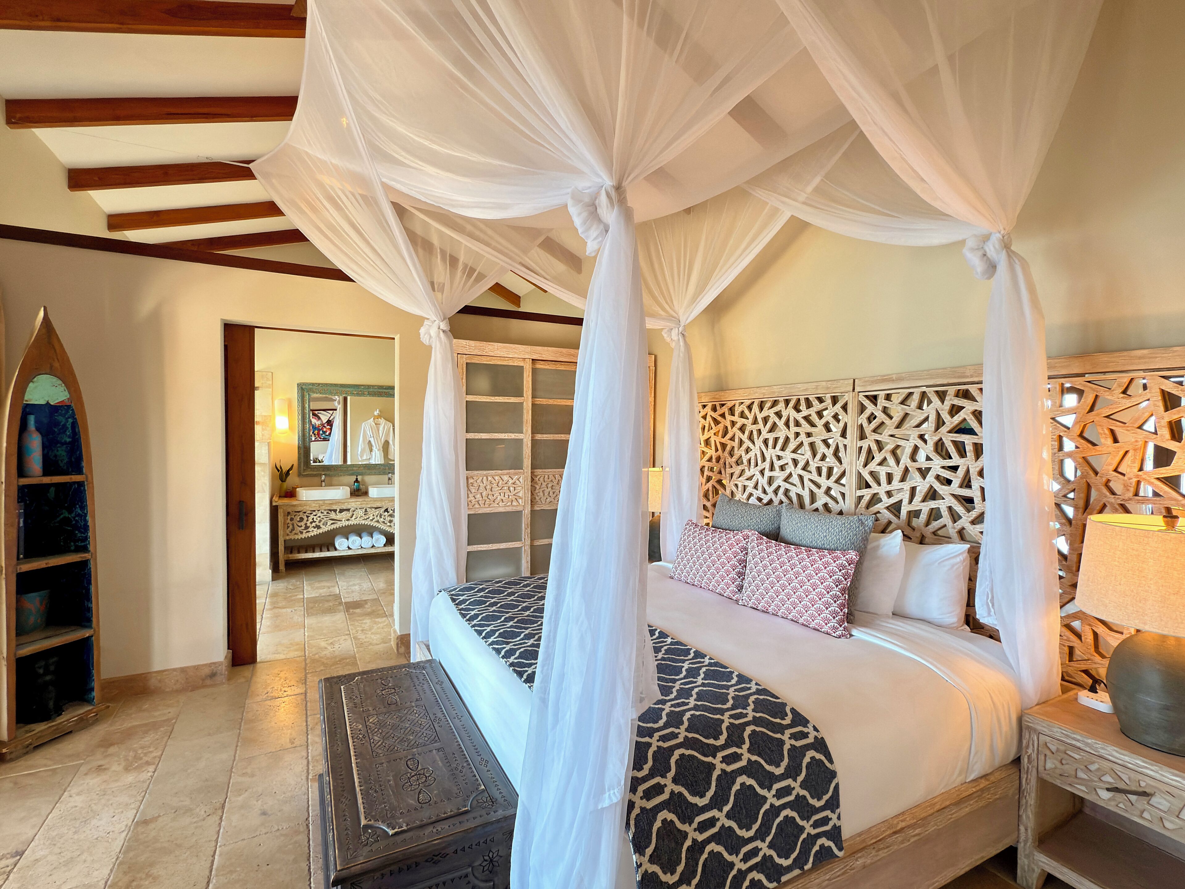 A bedroom with a canopy bed, a wooden headboard, a nightstand with a lamp, and a decorative wall panel.
