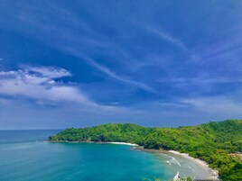 Paisagem costeira com praia, colinas arborizadas e céu azul límpido.