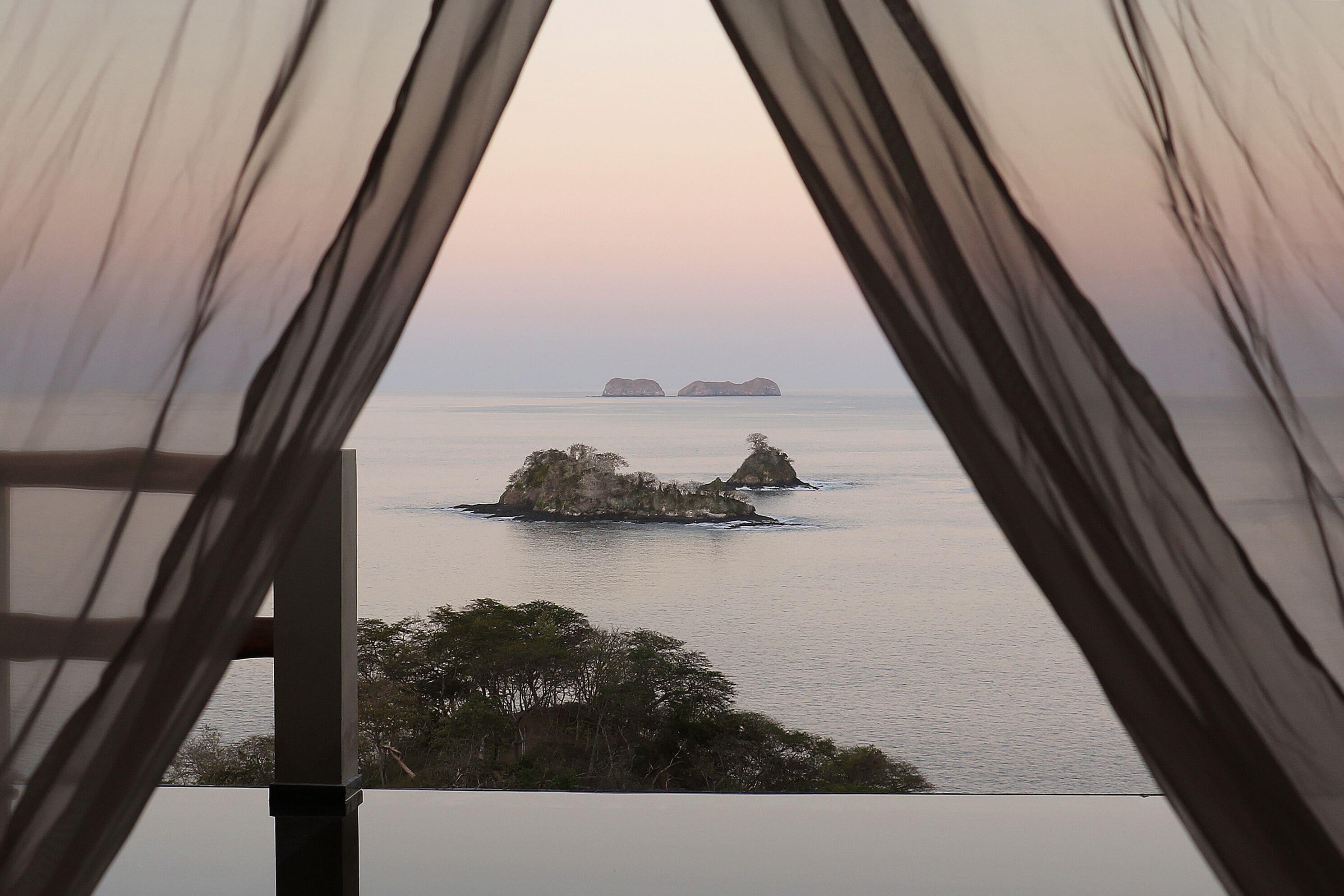 A view through sheer curtains of a calm sea with two prominent rock formations.
