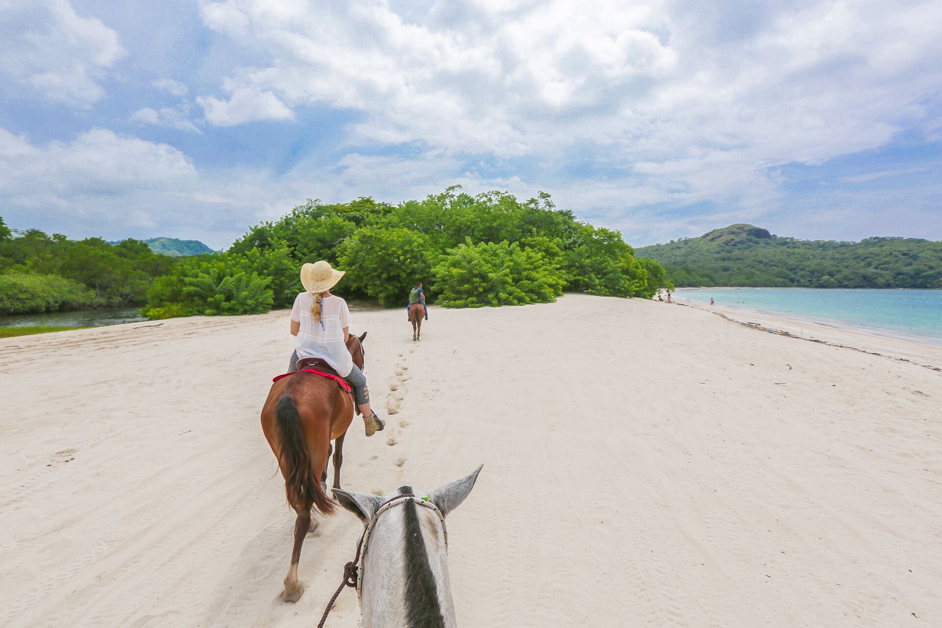 Uma praia com um cavalo e cavaleiro, céu claro e uma colina arborizada ao fundo.
