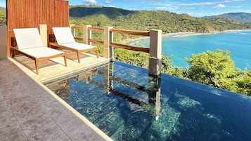 An infinity pool with two lounge chairs, a wooden deck, and a view of a beach and mountains.