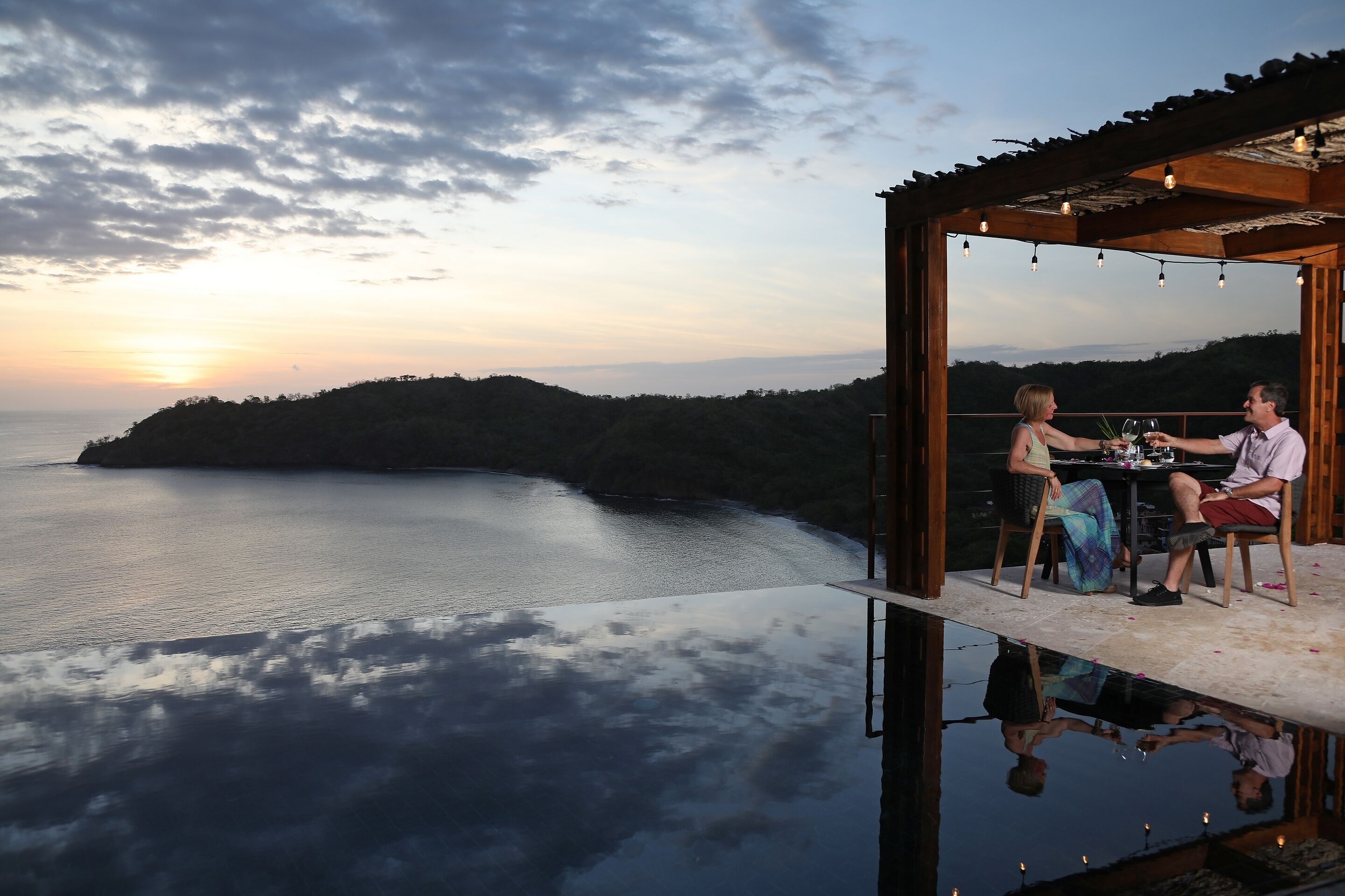 A couple dining on a glass floor balcony with a view of the sea and sunset.