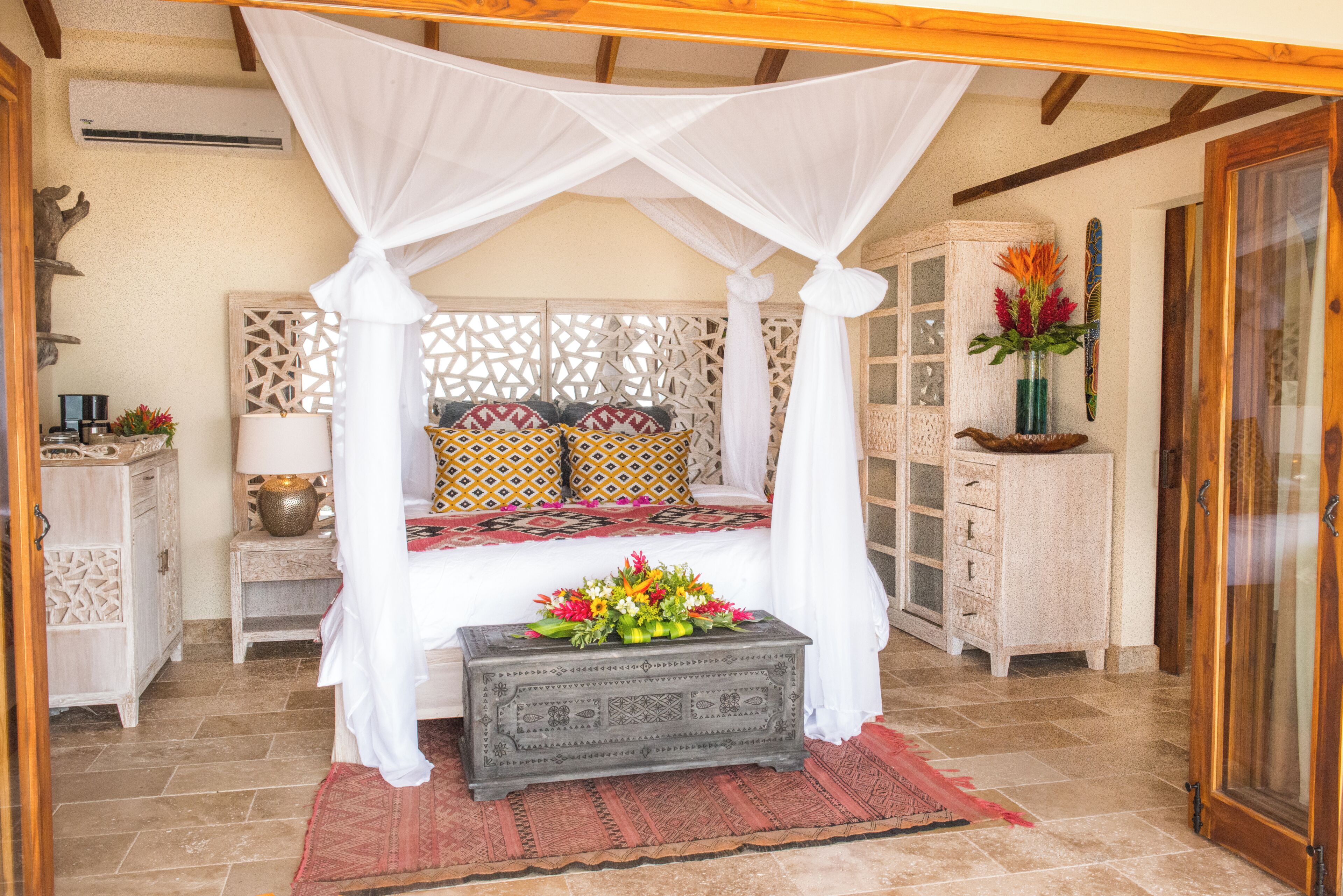 Une chambre avec un lit à baldaquin, une table de chevet avec une lampe, une petite table ornée d’un vase de fleurs et un meuble contenant des objets décoratifs.