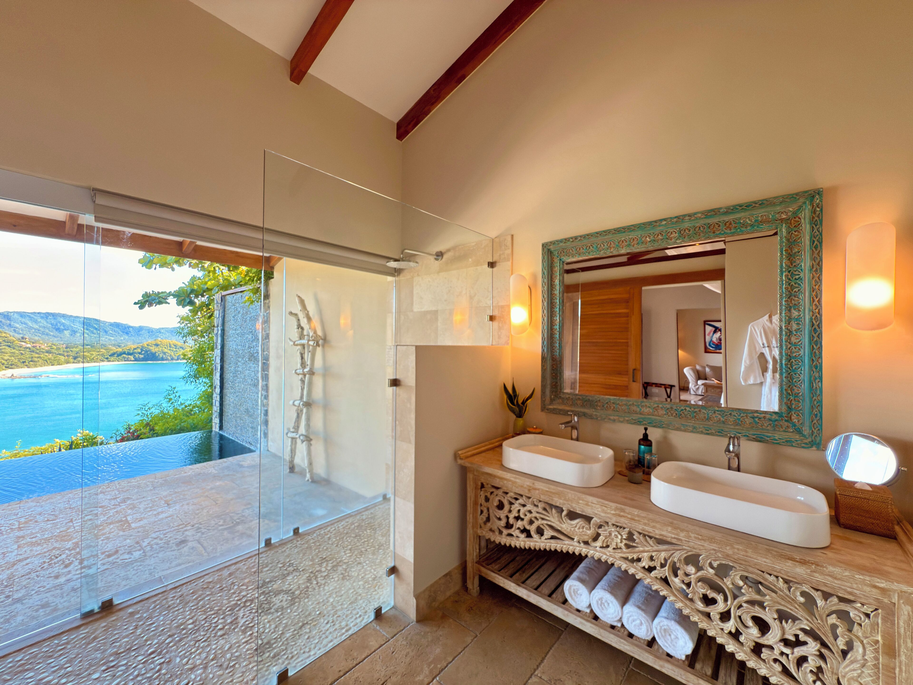 A bathroom with a large mirror, two sinks, a shower, and a view of a beach.