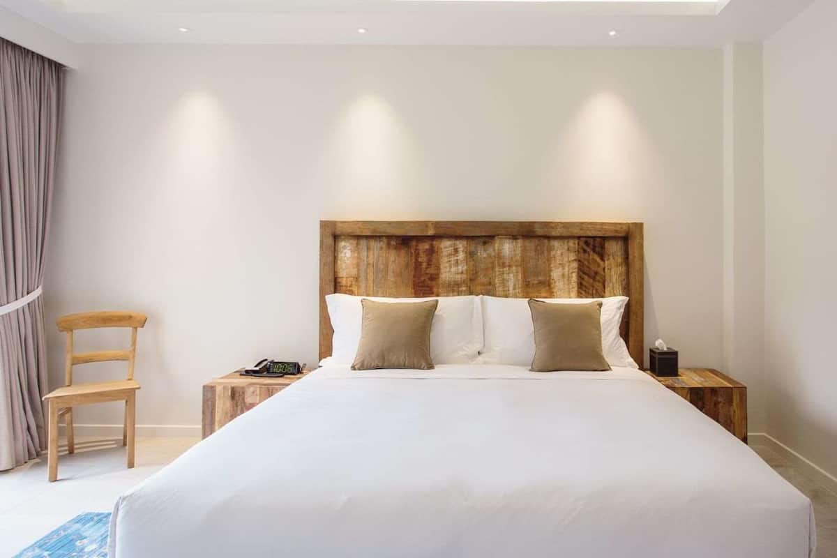 Quarto de hotel com uma cama grande, cabeceira de madeira, duas mesas de cabeceira, uma cadeira e um quadro na parede.