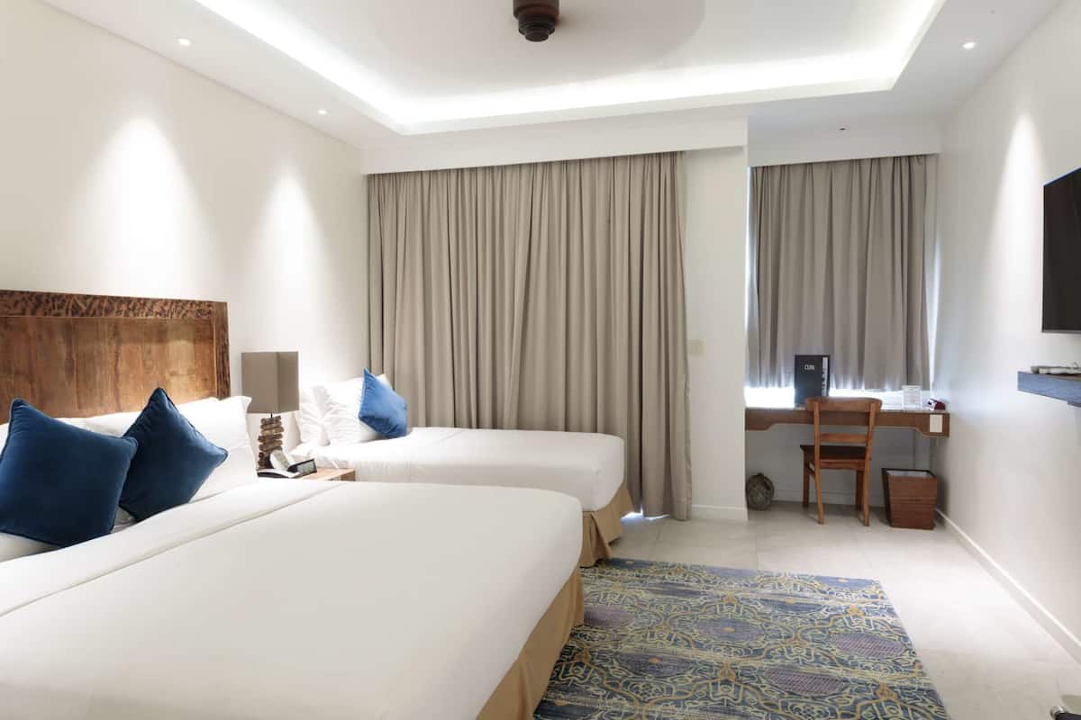 Quarto de hotel com duas camas, uma escrivaninha, uma cadeira, uma televisão e um tapete estampado.