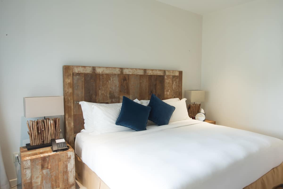 Uma cama bem arrumada com roupa de cama branca e duas almofadas azuis, uma cabeceira de madeira, uma luminária de cabeceira e um quadro na parede.
