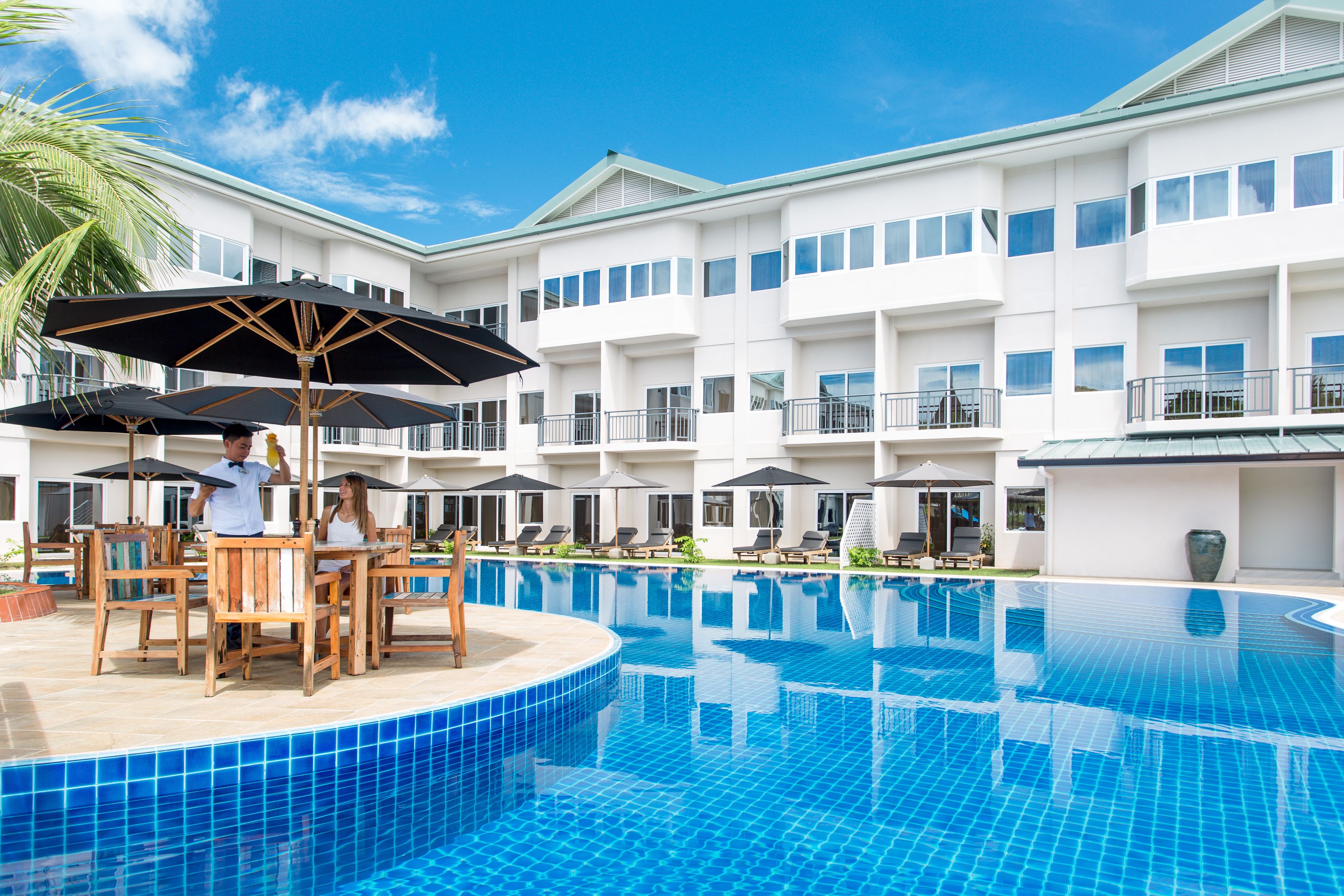 A hotel pool area with a white building, multiple umbrellas, and lounge chairs.