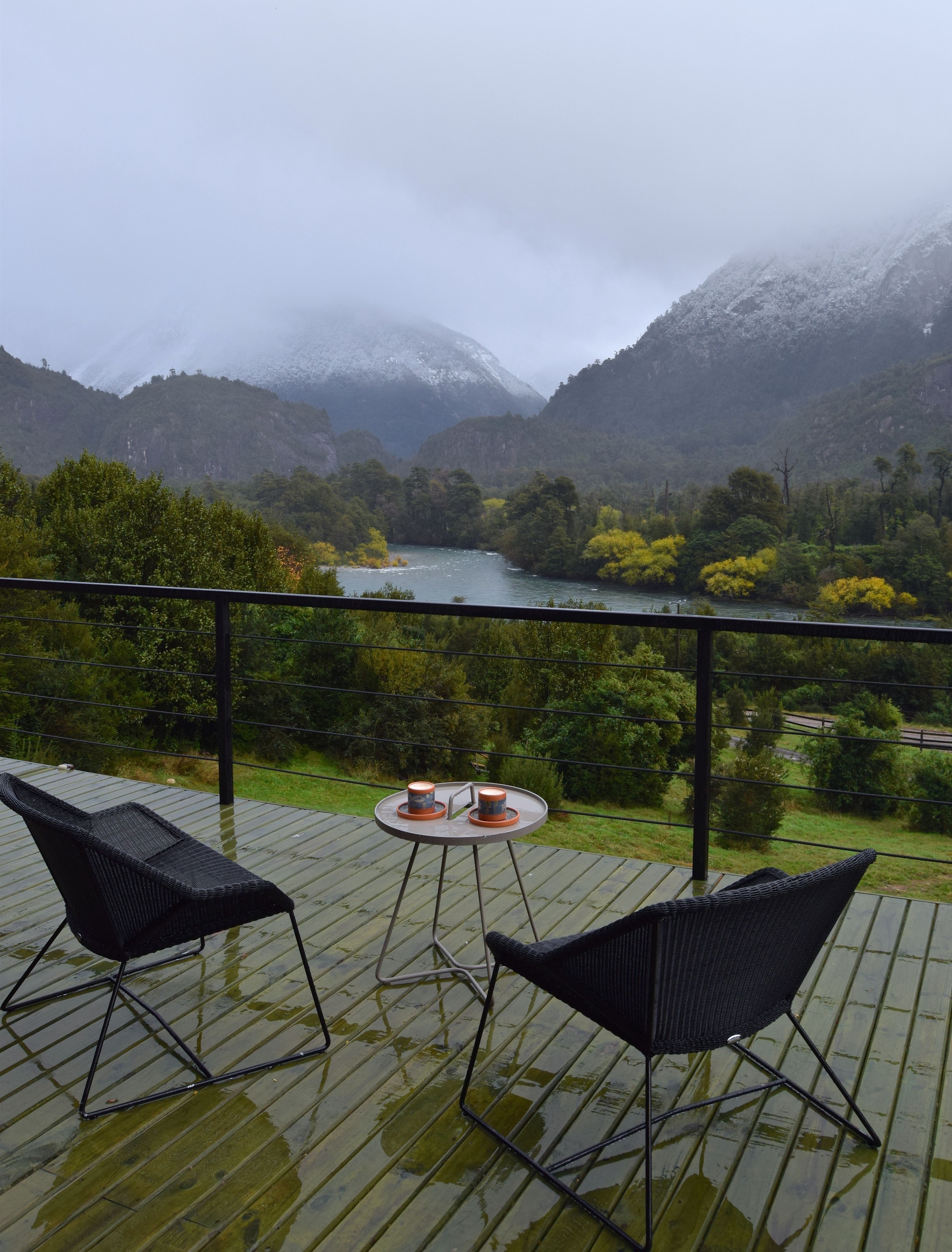 A deck with two chairs and a small table, overlooking a river and mountains.