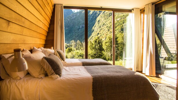 A bedroom with a large bed, two pillows, a bedside lamp, and a view of a mountainous landscape through a sliding glass door. - Hotel Raudal (Palena)