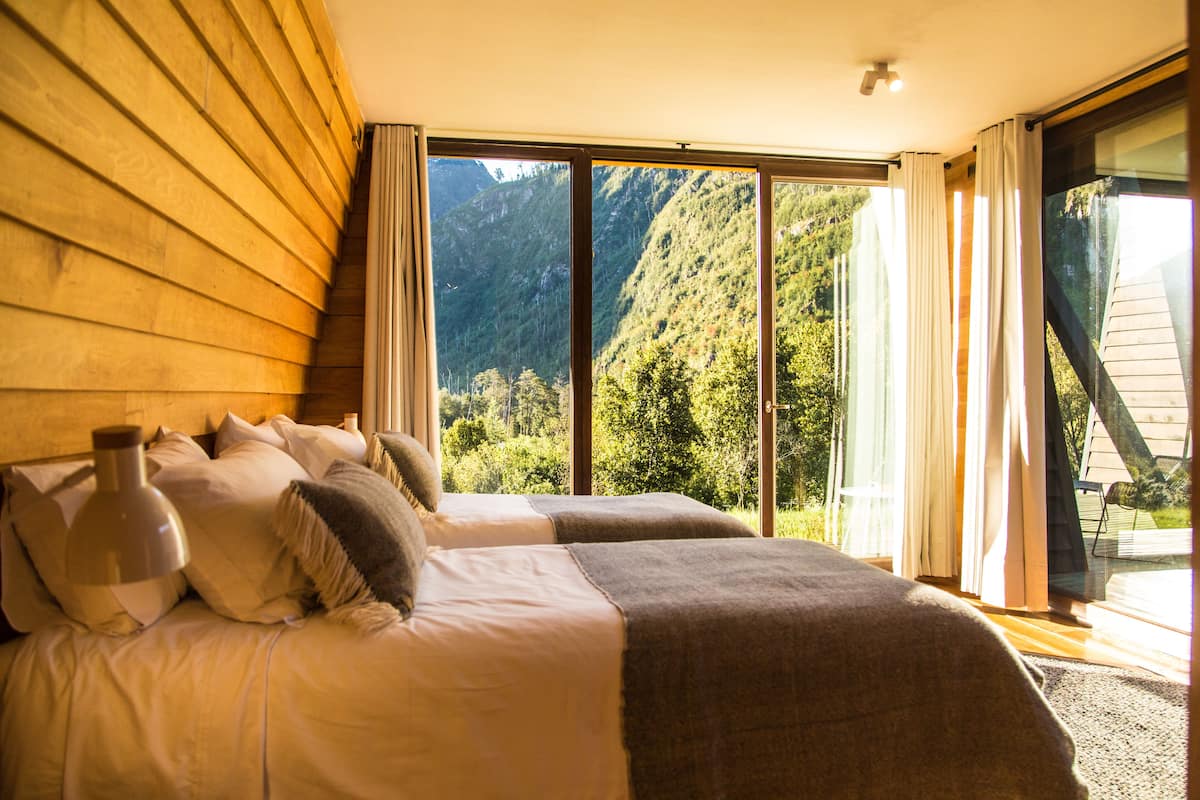 Un dormitorio con una cama grande, dos almohadas, una lámpara de noche y vistas a un paisaje montañoso a través de un ventanal corredero.