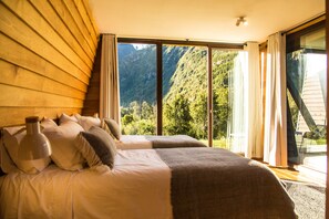 A bedroom with a large bed, two pillows, a bedside lamp, and a view of a mountainous landscape through a sliding glass door. - Hotel Raudal (Palena)