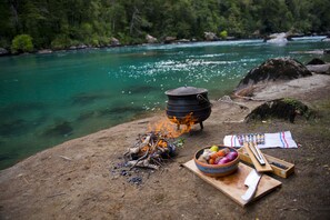 A campfire with cooking utensils and a bowl of food by a river. - Hotel Raudal (Palena)