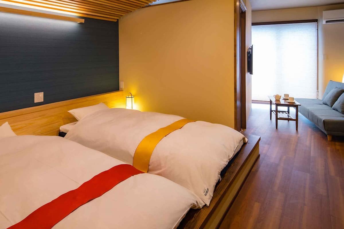 A hotel room with two beds, a wooden headboard, a small table with cups, and a sofa.