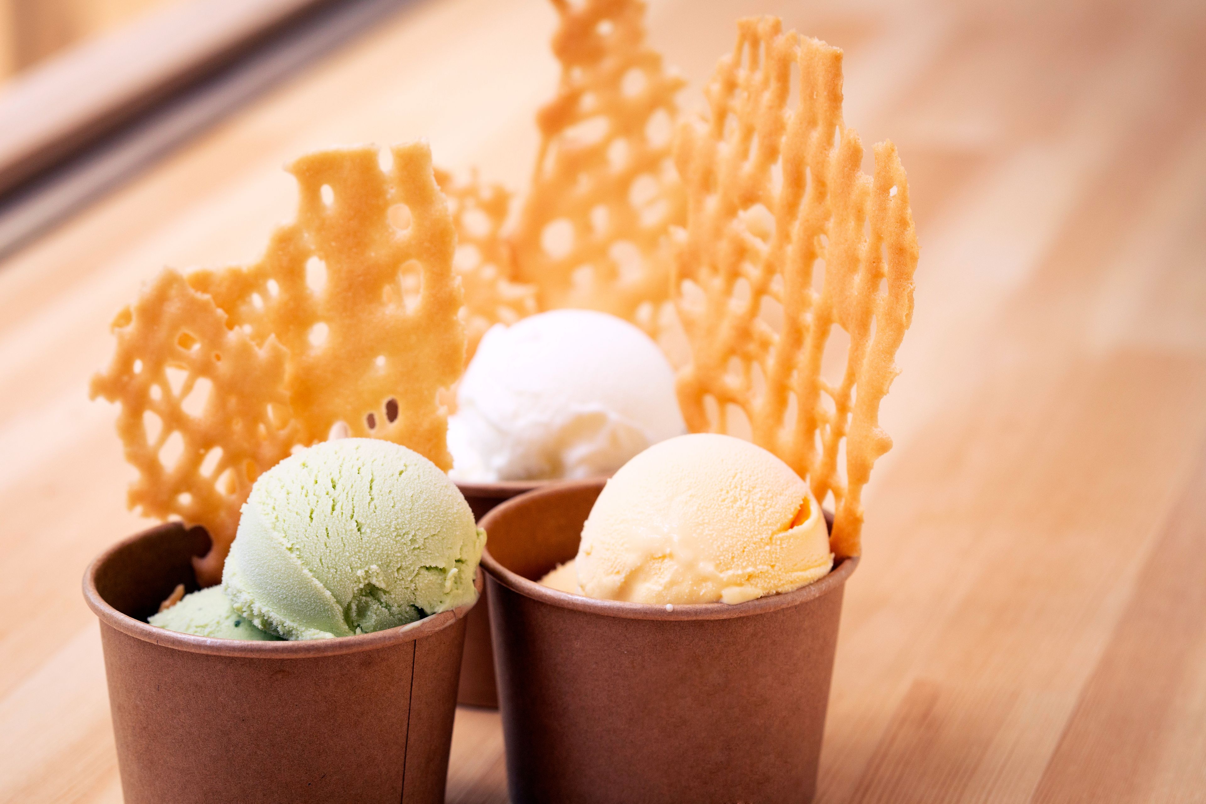 Three cups of ice cream in brown paper cups, each with a different flavor, and a decorative sugar sculpture in the background.
