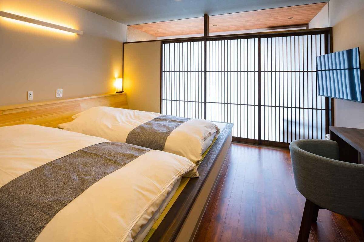 A hotel room with a bed, a chair, a wall-mounted light, and a large window with vertical blinds.