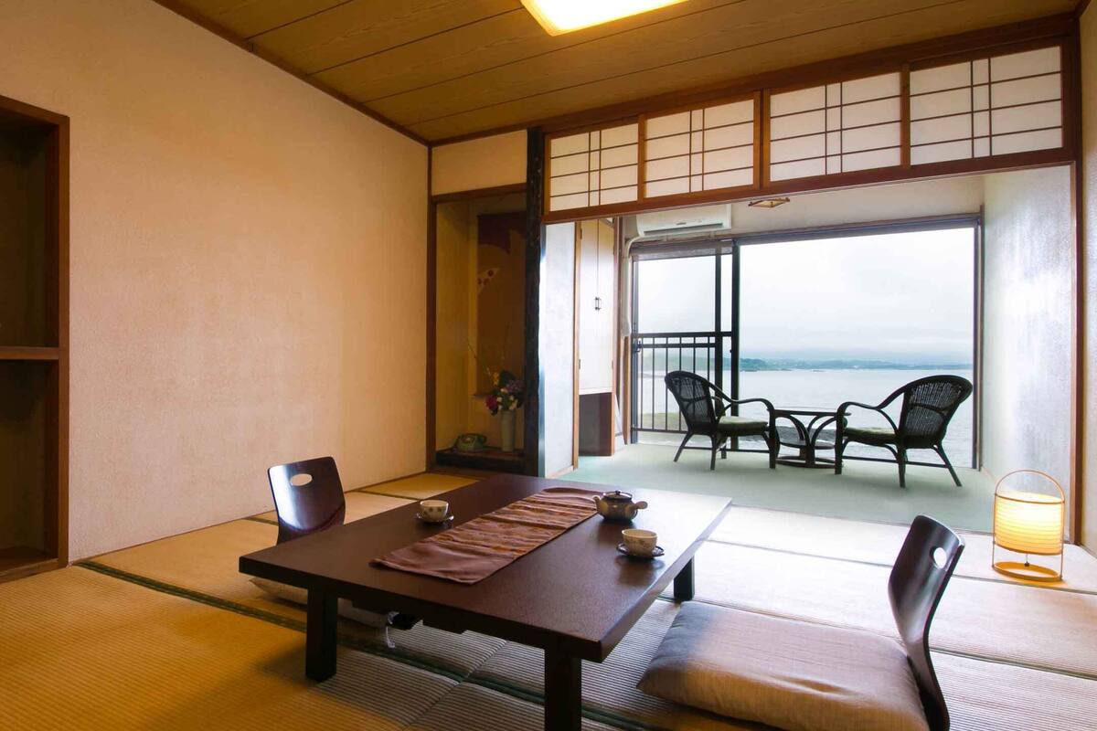 A traditional Japanese room with tatami flooring, a low table, and a view of the sea through large sliding doors.