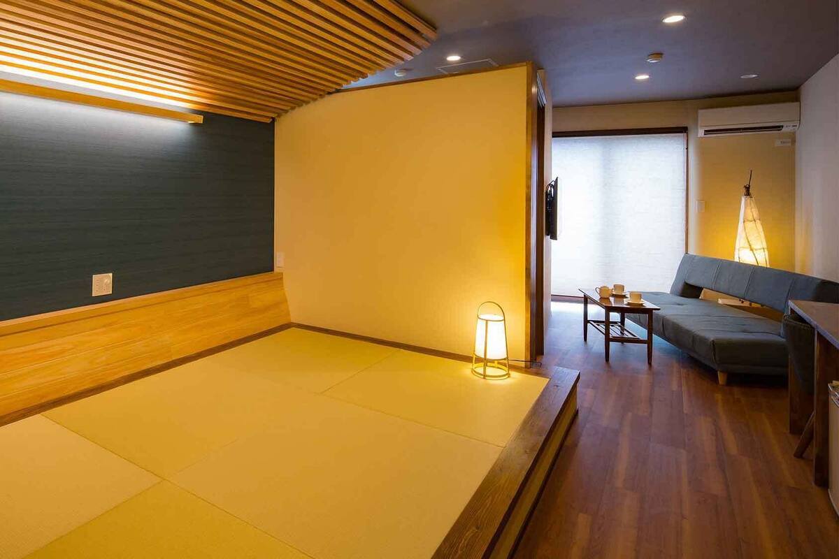A room with tatami flooring, a wooden ceiling, a sofa, a table with cups, and a lamp.
