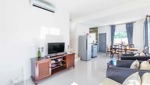 A modern living room with a flat-screen TV, a wooden entertainment unit, a dining area with a table and chairs, and a kitchen area with a refrigerator and a microwave.