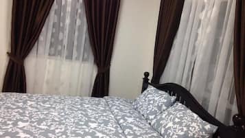A bed with a floral-patterned bedspread and matching pillows, dark wooden headboard, and sheer curtains with a floral pattern.
