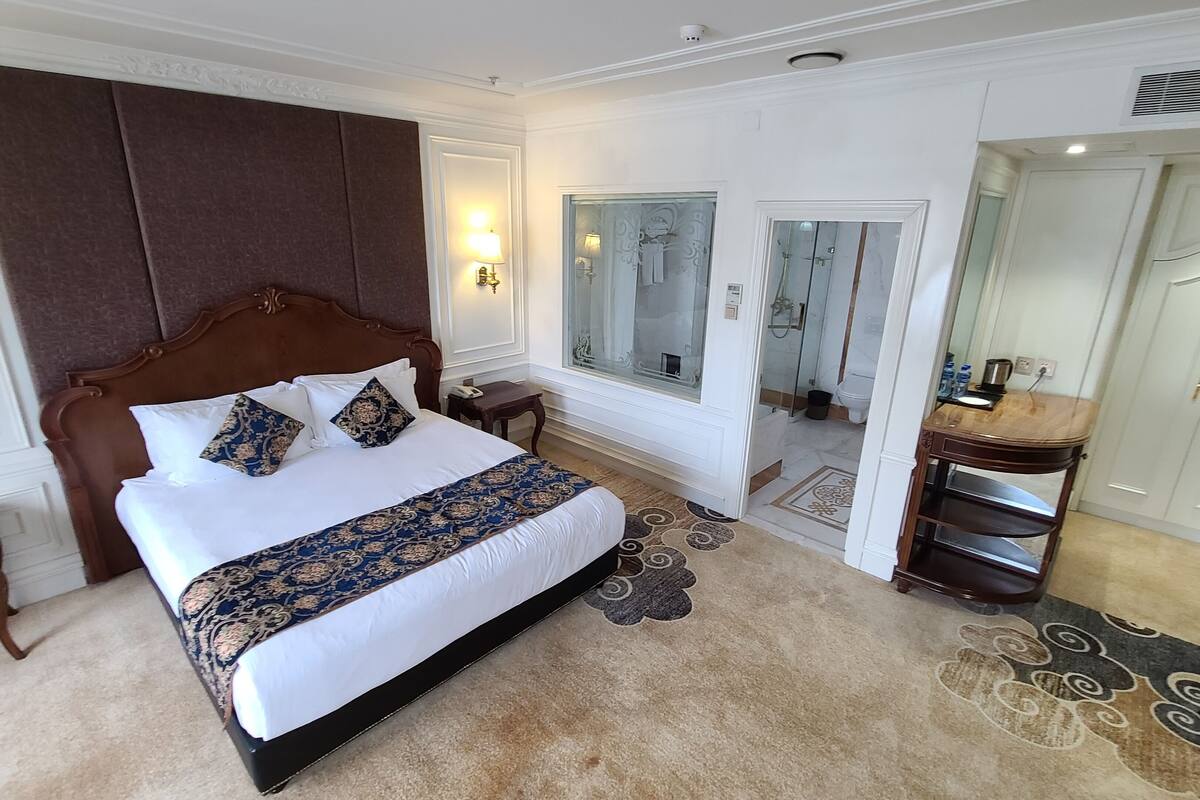 A large bed with a headboard, bedside tables, a nightstand, a mirror, and a bathroom visible through a glass door.
