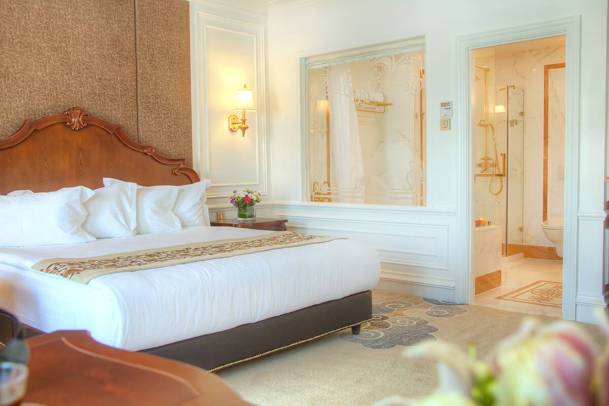 A hotel room with a large bed, a wooden headboard, a nightstand with a vase of flowers, and a bathroom with a glass shower enclosure.