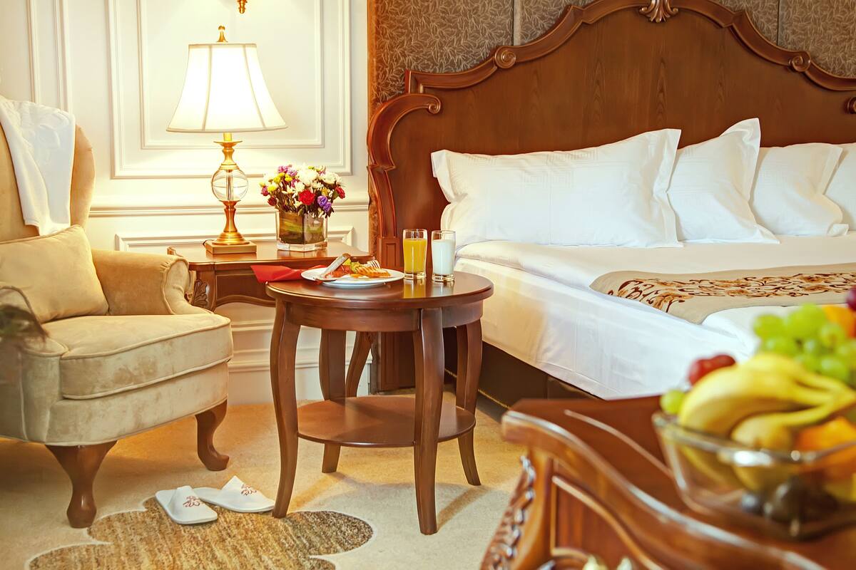 A hotel room with a bed, a chair, a table with a lamp, a vase of flowers, and a bowl of fruit.