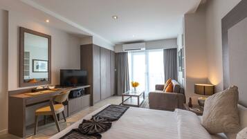 A modern hotel room with a large bed, a desk with a computer, a TV, and a seating area with a sofa and armchair.