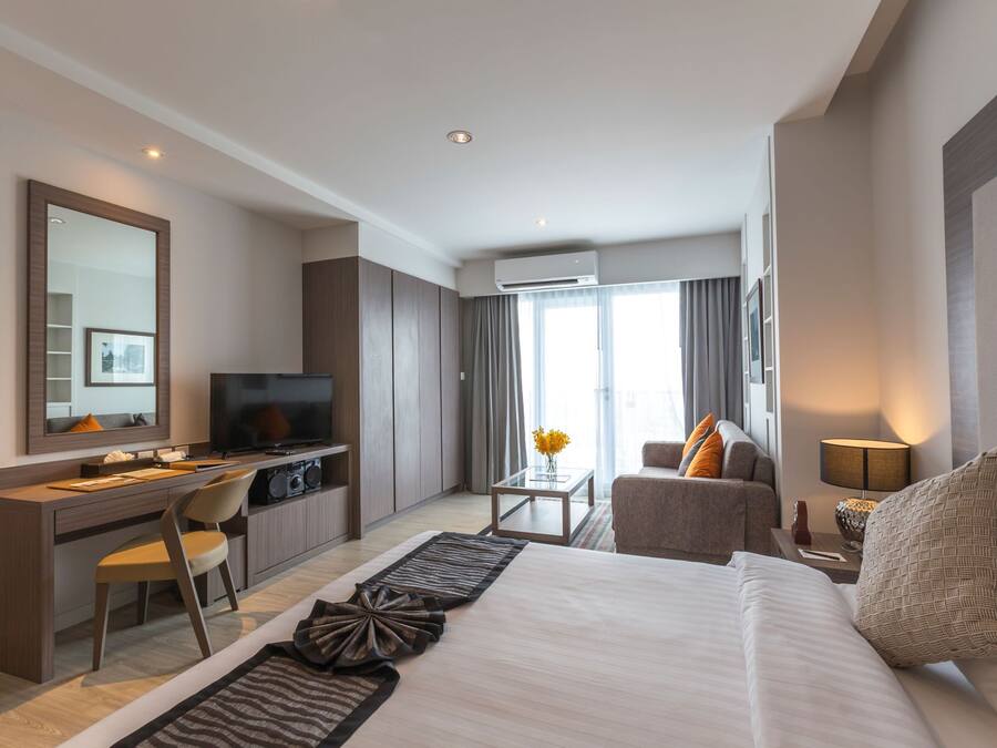 A modern hotel room with a large bed, a desk with a computer, a TV, and a seating area with a sofa and armchair.