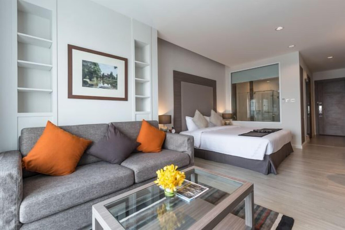 Ett modernt hotellrum med en grå soffa, en säng med vita sängkläder, ett glasbord med en vas med blommor och en inramad tavla på väggen.