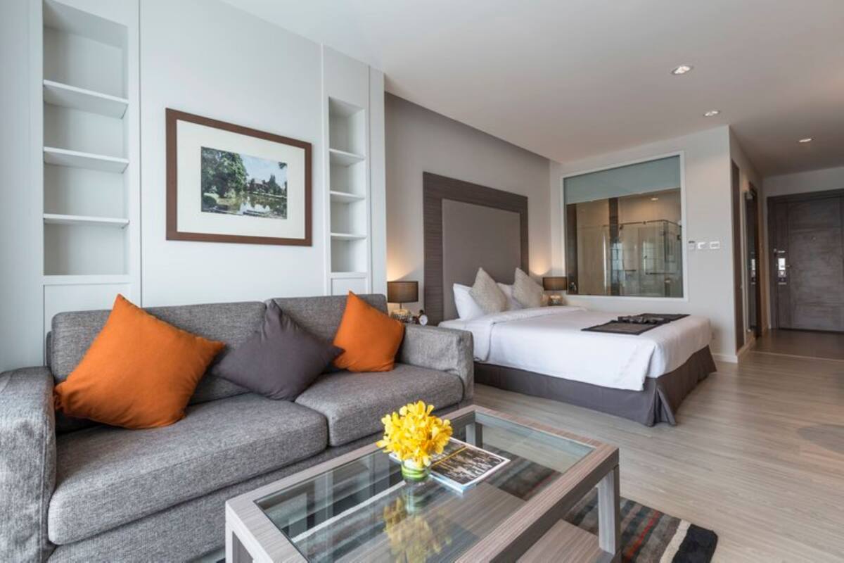A modern hotel room with a grey sofa, a bed with white bedding, a glass coffee table with a magazine and flowers, and a framed picture on the wall.