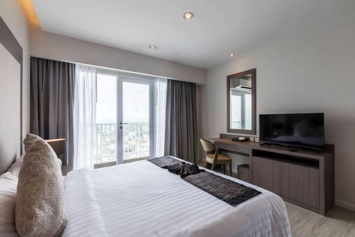 A hotel room with a large bed, a desk, a television, and a balcony with curtains.
