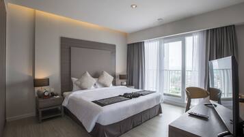 A modern hotel room with a large bed, a desk with a computer, and a view of the city through large windows.