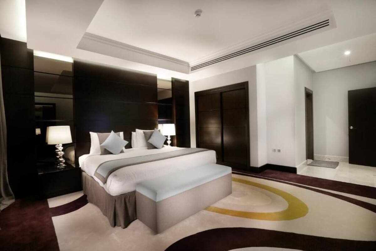A modern hotel room with a large bed, dark wood paneling, a nightstand with a lamp, and a large mirror.