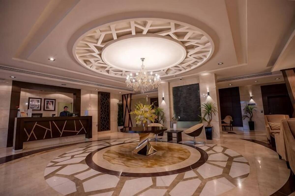 A hotel lobby with a reception desk, a chandelier, a circular patterned floor, and a large plant in the center.
