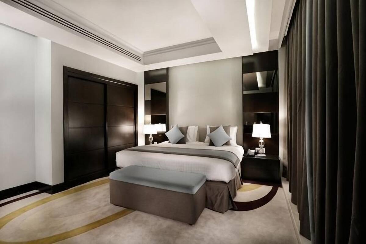 A modern hotel room with a large bed, a bedside table with a lamp, a dark wardrobe, and a large window with curtains.