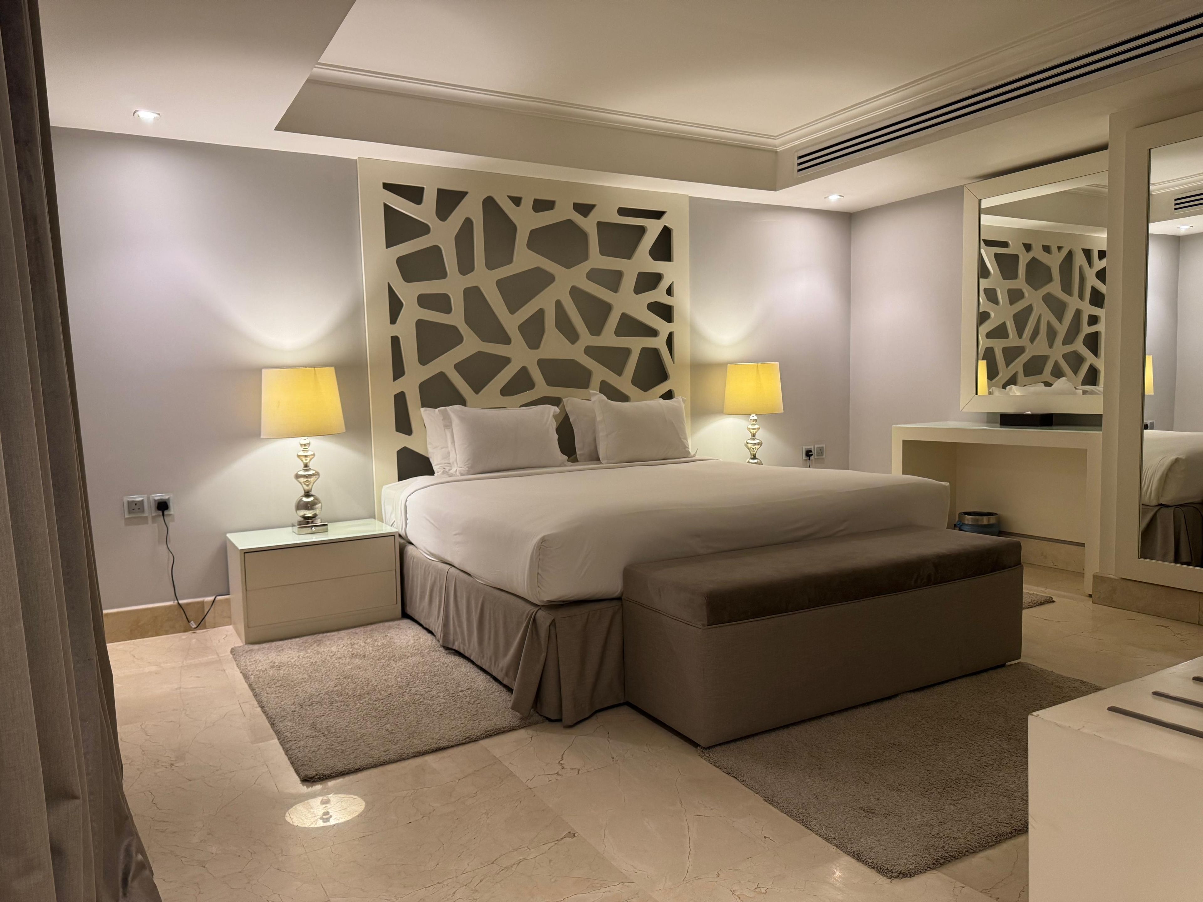 Una habitación de hotel moderna con una cama grande, lámparas de noche, un adorno empotrado en la pared, una mesita de noche y un espejo.