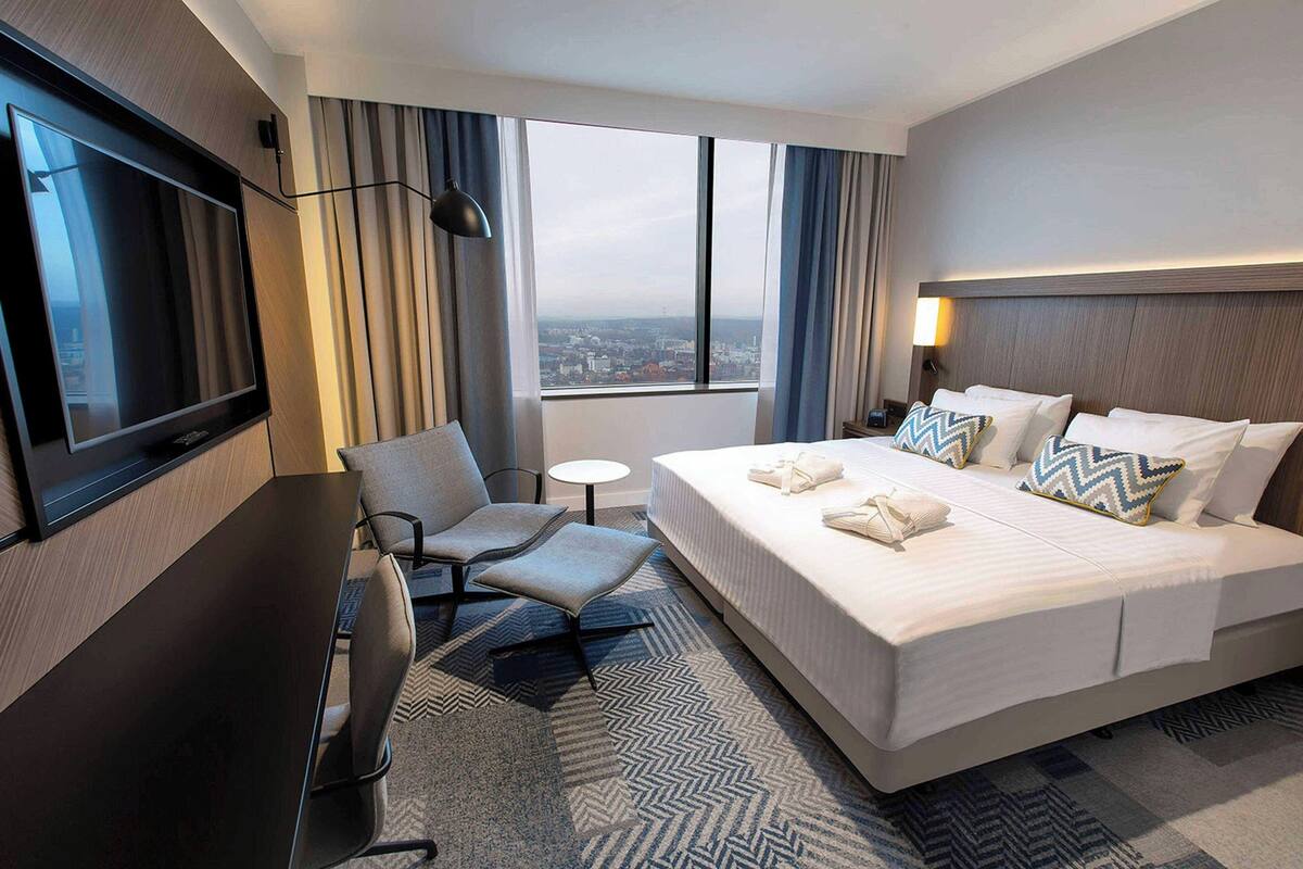 Une chambre d’hôtel avec un grand lit, un bureau, une chaise, une télévision et une vue sur la ville par la fenêtre.