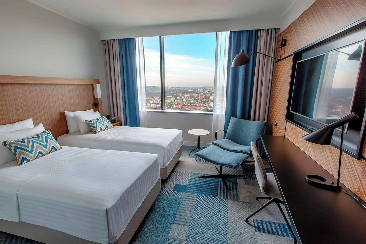 Une chambre d’hôtel avec deux lits, un bureau avec une télévision, une chaise, une petite table et une vue sur la ville grâce à de grandes fenêtres.