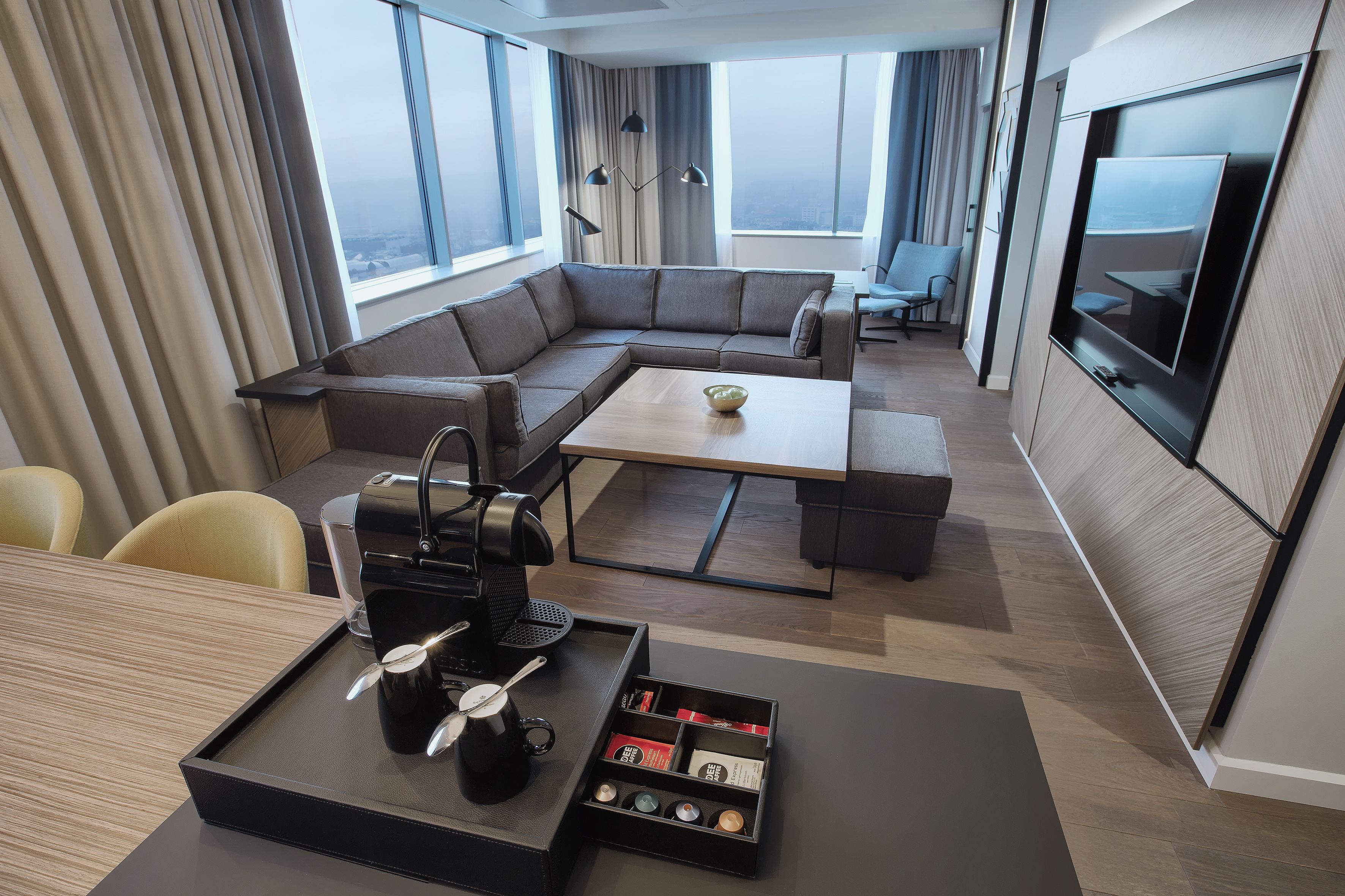 A modern hotel room with a sectional sofa, a coffee table, a TV, and a dining area.