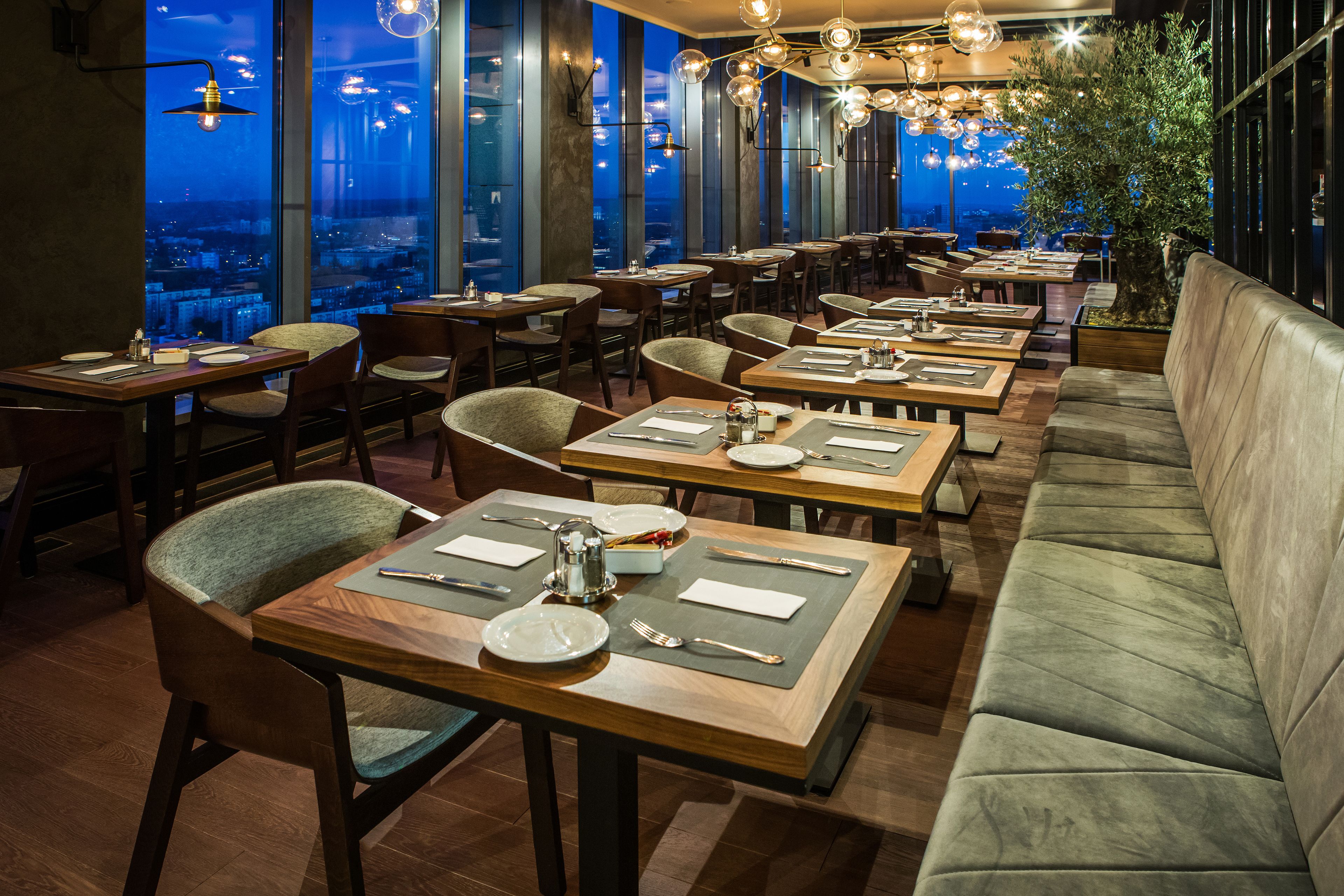 A restaurant with multiple tables and chairs, large windows offering a city view, and a seating area with a sofa.