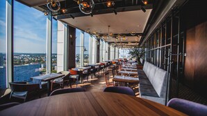 Un restaurant avec de hauts plafonds, de grandes fenêtres et une vue sur la ville. Les tables et les chaises sont disposées pour accueillir les clients.