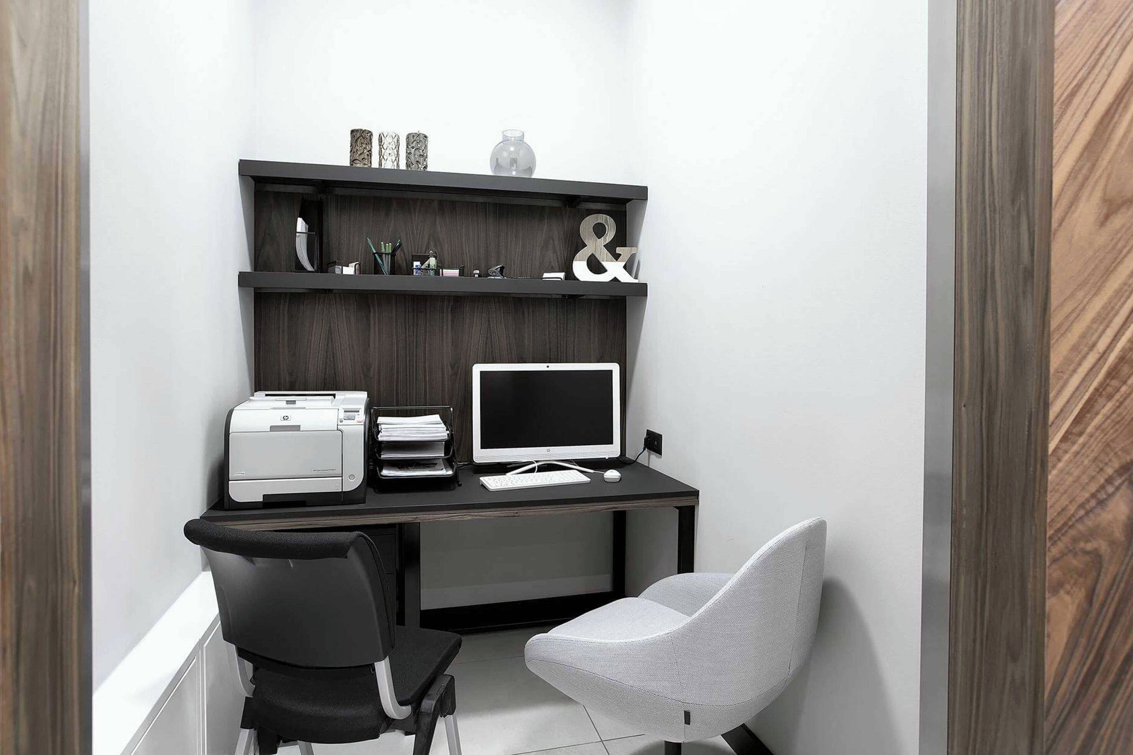 A compact office space with a desk, two chairs, a printer, a monitor, and a shelf with decorative items.