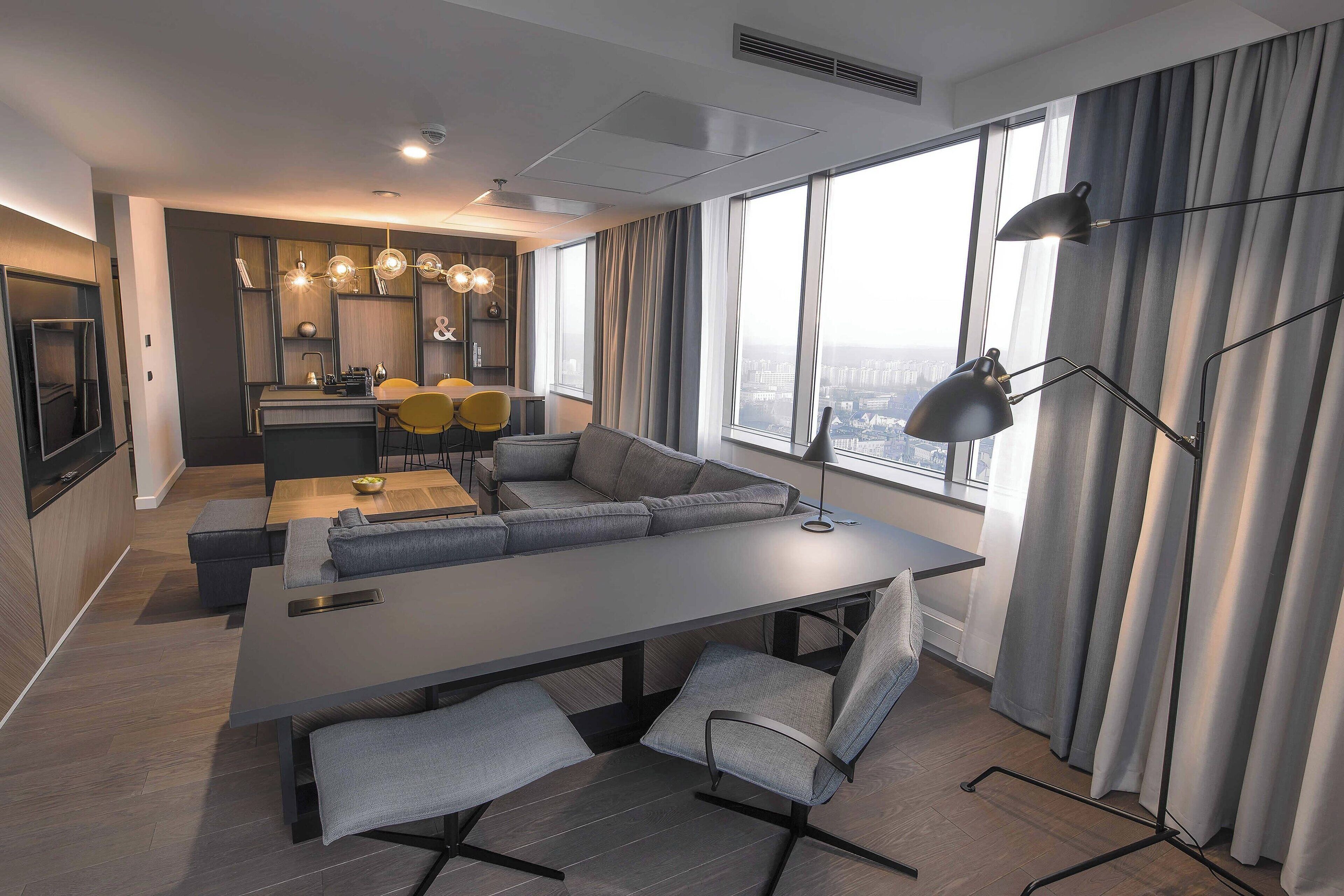 A modern hotel room with a large dining table, grey sofas, and a city view.
