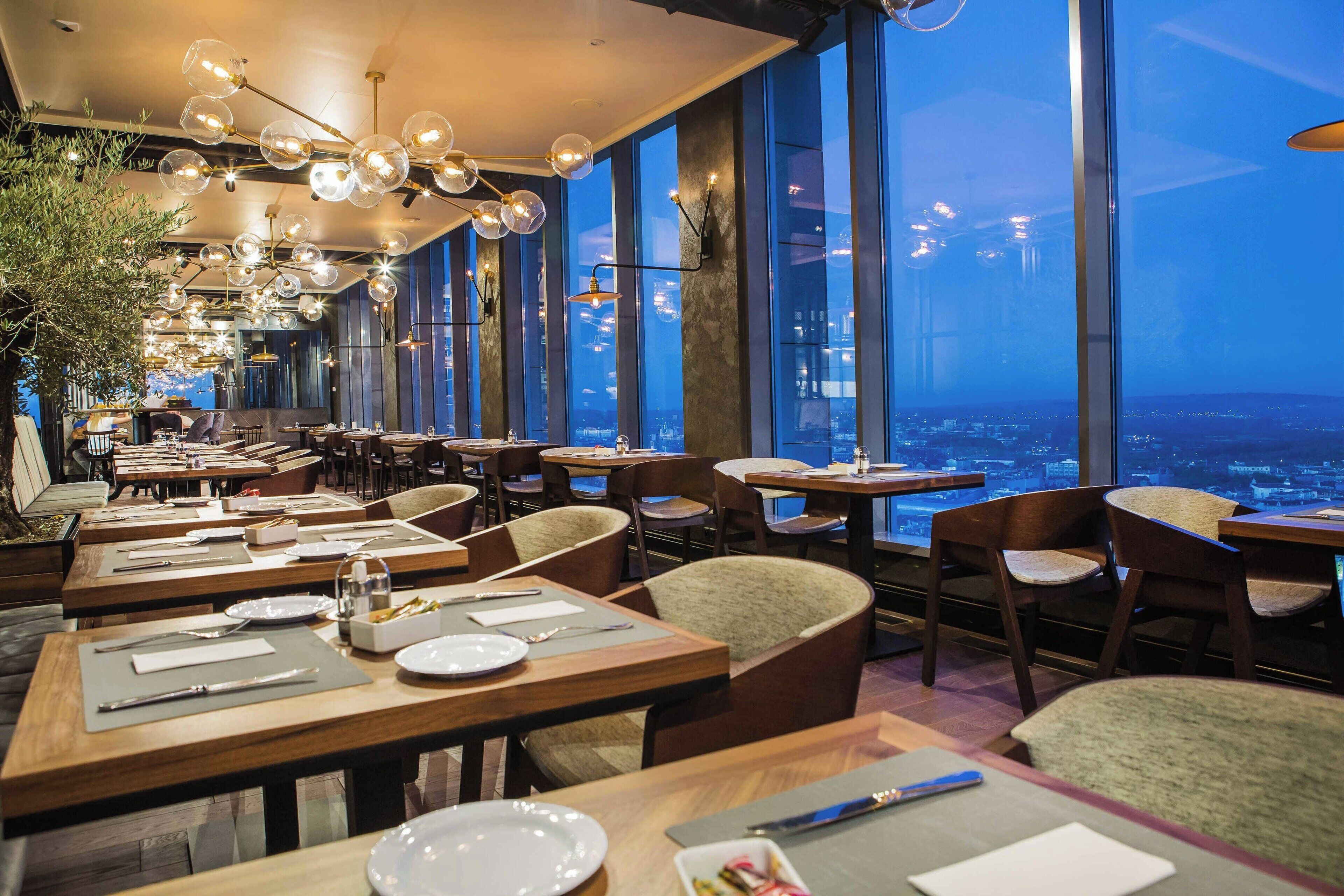 A restaurant with tables set for dining, large windows offering a city view, and hanging spherical light fixtures.